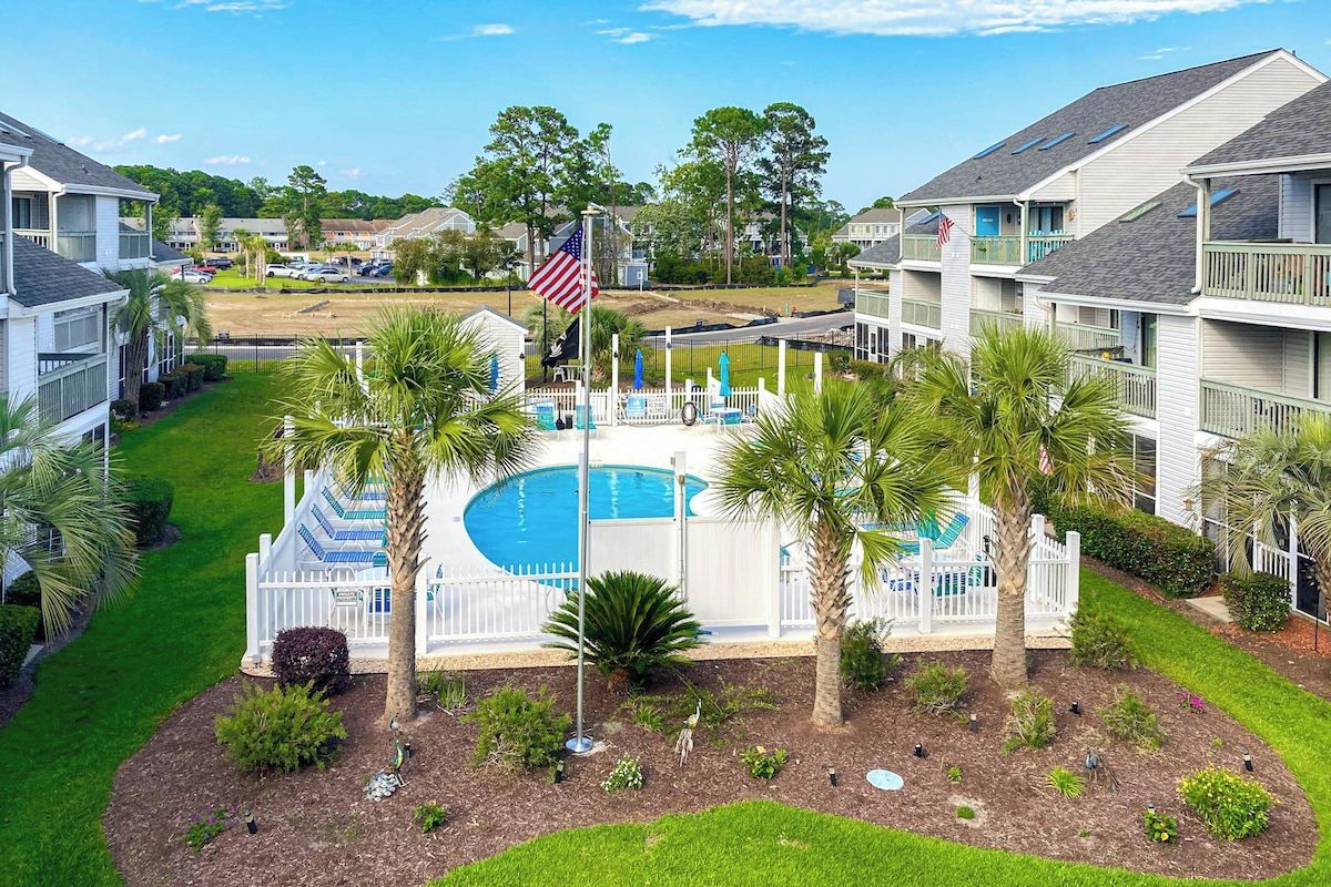A residential complex with a swimming pool surrounded by palm trees and a white picket fence.