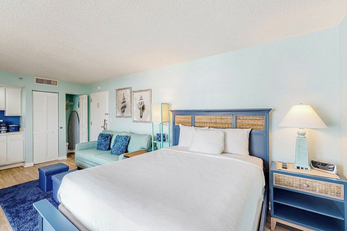 A bedroom with a large bed, a nightstand with a lamp, a wardrobe, and a small seating area with a sofa.