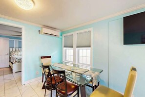 A dining area with a glass table and chairs, a window with blinds, and an air conditioning unit on the wall. - Flamingo House 1 - bottom (North Myrtle Beach)