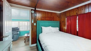 Un dormitorio con techo de madera, cama con ropa de cama blanca, cabecera verde azulado y cortina roja.