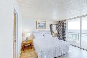 A bedroom with a large bed, a nightstand, a lamp, a framed picture on the wall, and a view of the sea through a balcony. - Water's Edge Luxe | Nine Oh One (Murrells Inlet)