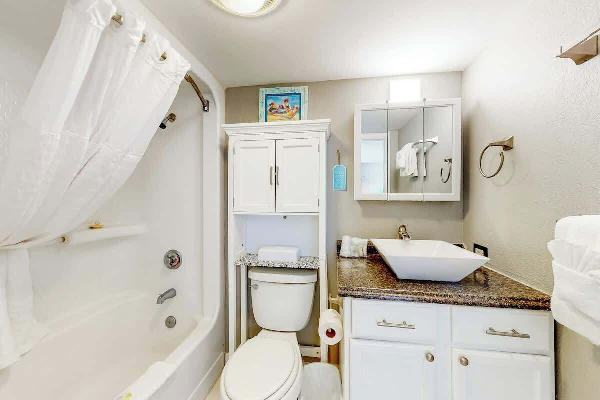 A bathroom with a white bathtub, a toilet, a sink with a granite countertop, and a cabinet with drawers.