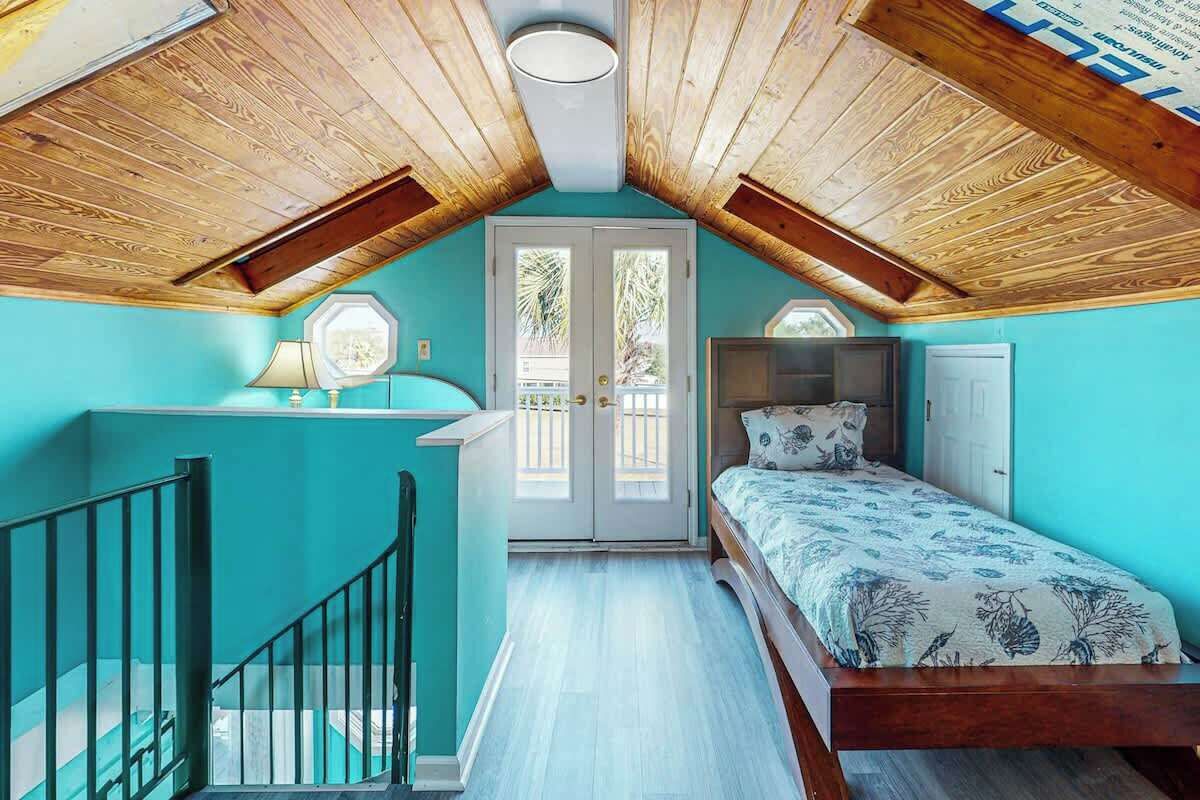 Une chambre avec un plafond en pente, un lit recouvert d’une literie à motifs floraux, une fenêtre donnant sur un beau paysage et un escalier.