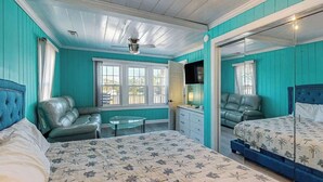 A room with teal walls, a bed with floral bedding, a sofa, a television, and a ceiling fan. - Flamingo House 2 - top (North Myrtle Beach)