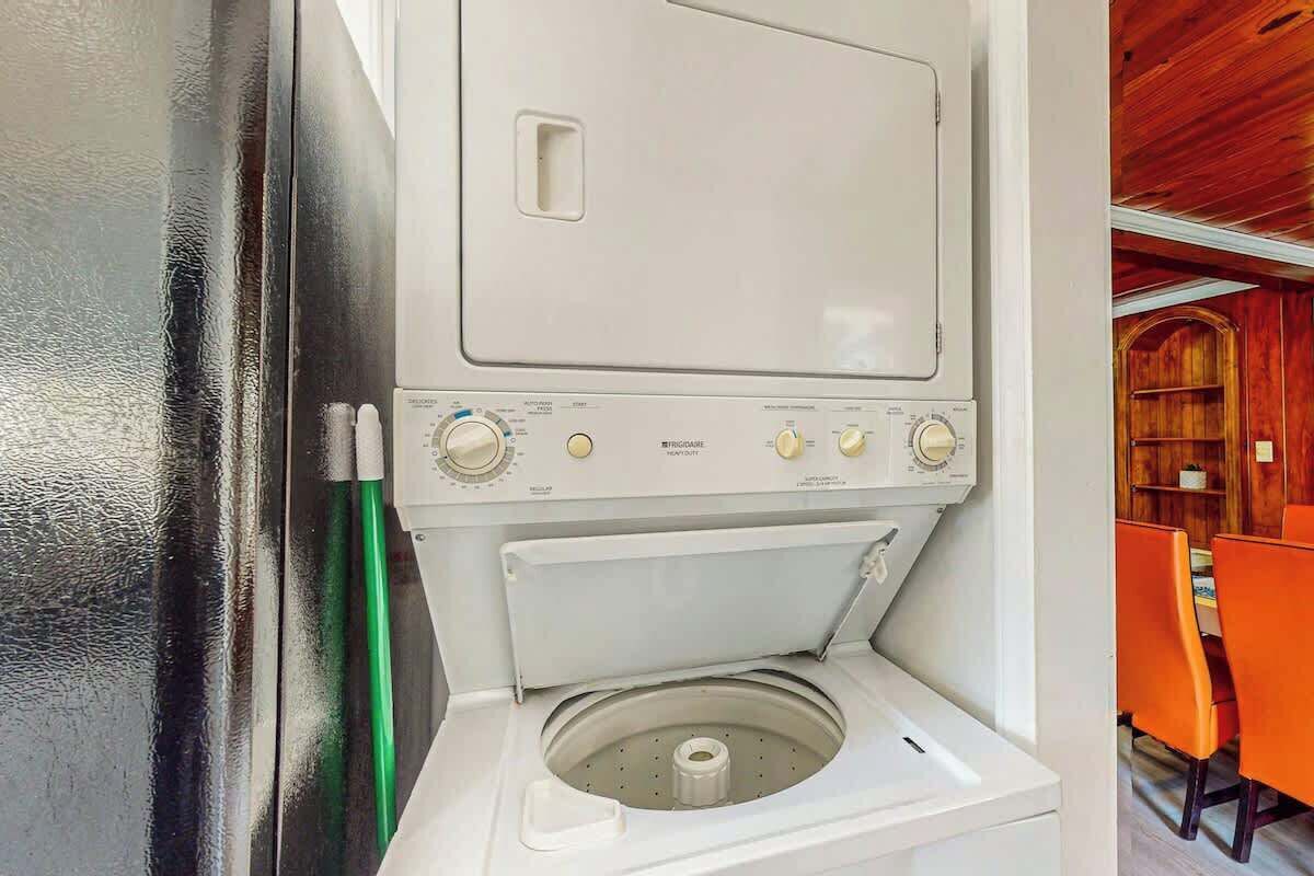 Lavadora blanca y compacta con perillas de control y puerta frontal, ubicada en una habitación con paredes de madera y sillas color naranja.