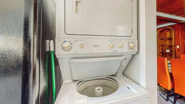 A compact, white laundry machine with control knobs and a front-loading door, located in a room with wooden walls and orange chairs. - Flamingo House 2 - top (North Myrtle Beach)