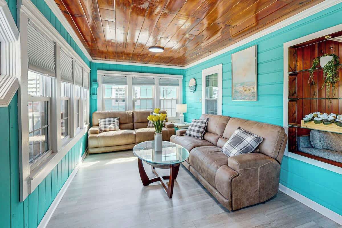 A room with a wooden ceiling, teal walls, a brown leather sofa, a glass coffee table, and a window with blinds.