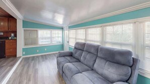 Un salon avec un canapé gris, des murs de couleur turquoise et de grandes fenêtres équipées de stores.