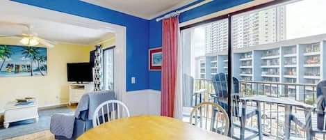 A room with a dining table, chairs, a ceiling fan, a TV, and a view of a building through a large window.