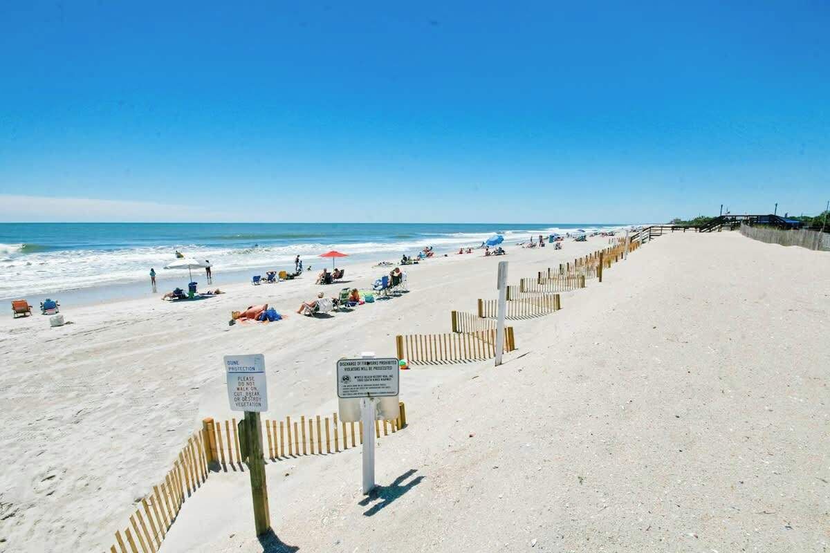 A sandy beach with a wooden fence, beachgoers, and a clear blue sky.