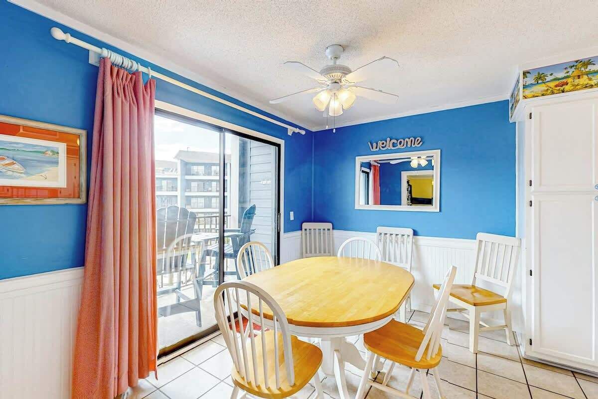 Una habitación con pared azul, una mesa redonda de madera con sillas, un ventilador de techo y una ventana con vistas a edificios.