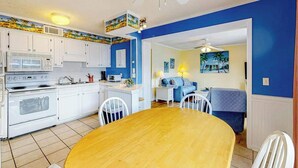A compact kitchen with white cabinets, a microwave, and a stove. There is a dining area with a wooden table and chairs. The walls are painted blue, and there is a ceiling fan. - Myrtle Beach Resort A-332 (Myrtle Beach)