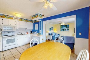 A compact kitchen with white cabinets, a microwave, and a stove. There is a dining area with a wooden table and chairs. The walls are painted blue, and there is a ceiling fan. - Myrtle Beach Resort A-332 (Myrtle Beach)