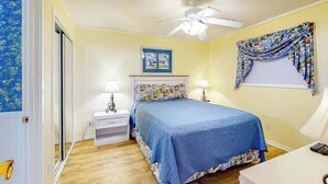 A bedroom with a bed, two bedside lamps, a ceiling fan, a window with curtains, and a door with a blue floral pattern. - Myrtle Beach Resort A-332 (Myrtle Beach)