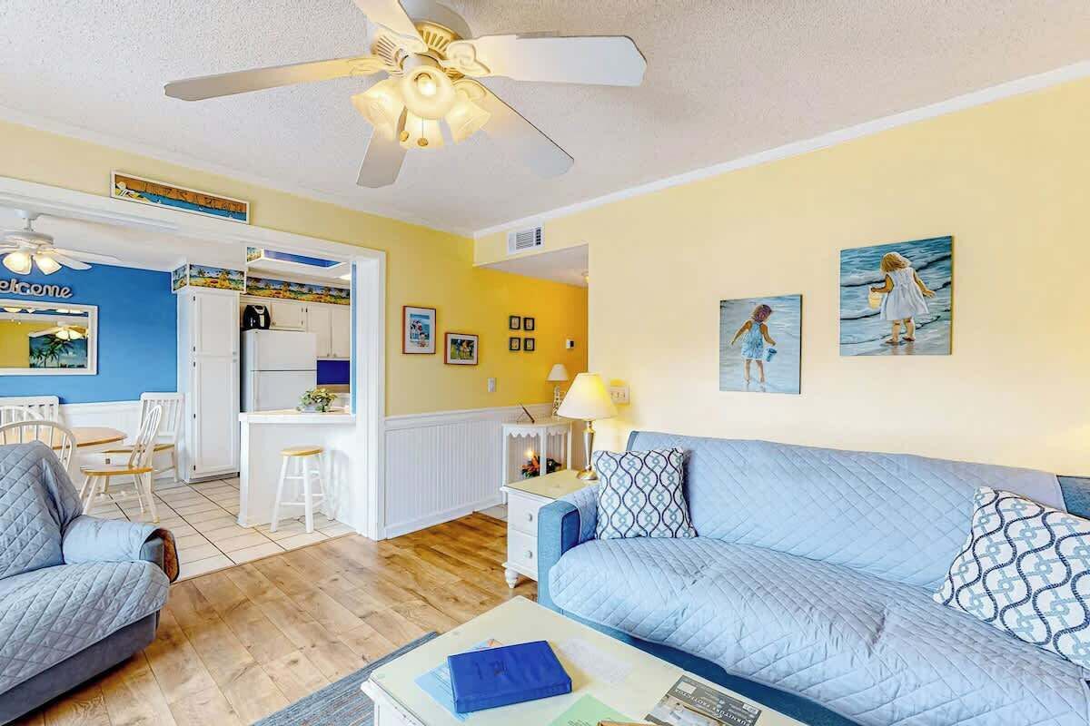 Ein Wohnzimmer mit blauem Sofa, einem Couchtisch und einem Deckenventilator. Es gibt eine Küchenzeile mit weißen Schränken und einem Esstisch.