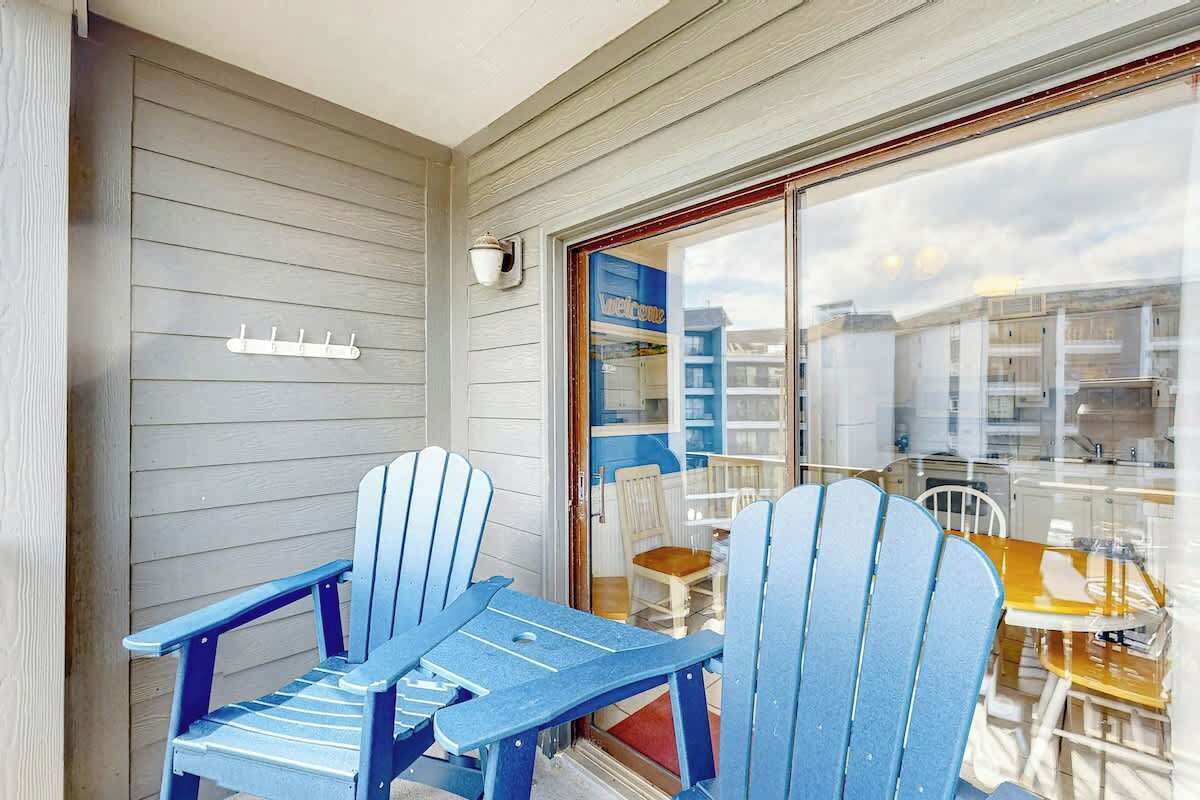 A balcony with two blue Adirondack chairs and a table, overlooking a residential area with houses and a clear sky.