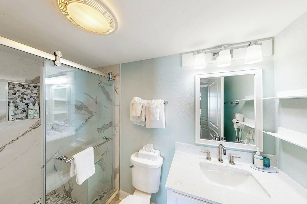 Ein modernes Badezimmer mit einer Duschkabine aus Glas, einem weißen Waschbecken mit großem Spiegel und einer Toilette mit Toilettenpapierhalter.