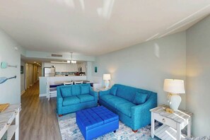 A living room with a blue sectional sofa, a matching ottoman, and a white side table with a lamp. - Bay Watch Resort 0315 (North Myrtle Beach)
