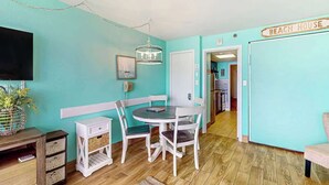 Een kamer met een blauwgroene muur, een eettafel met stoelen, een televisie en een bordje met de tekst 'BEACH HOUSE'.