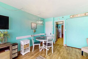 A room with a teal wall, a dining table with chairs, a television, and a sign that reads 'BEACH HOUSE'. - Sandcastle South #616 (Myrtle Beach)