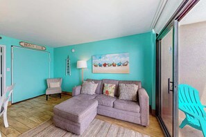 A room with a teal wall, a grey sofa, a white chair, a teal chair, and a painting of boats. - Sandcastle South #616 (Myrtle Beach)