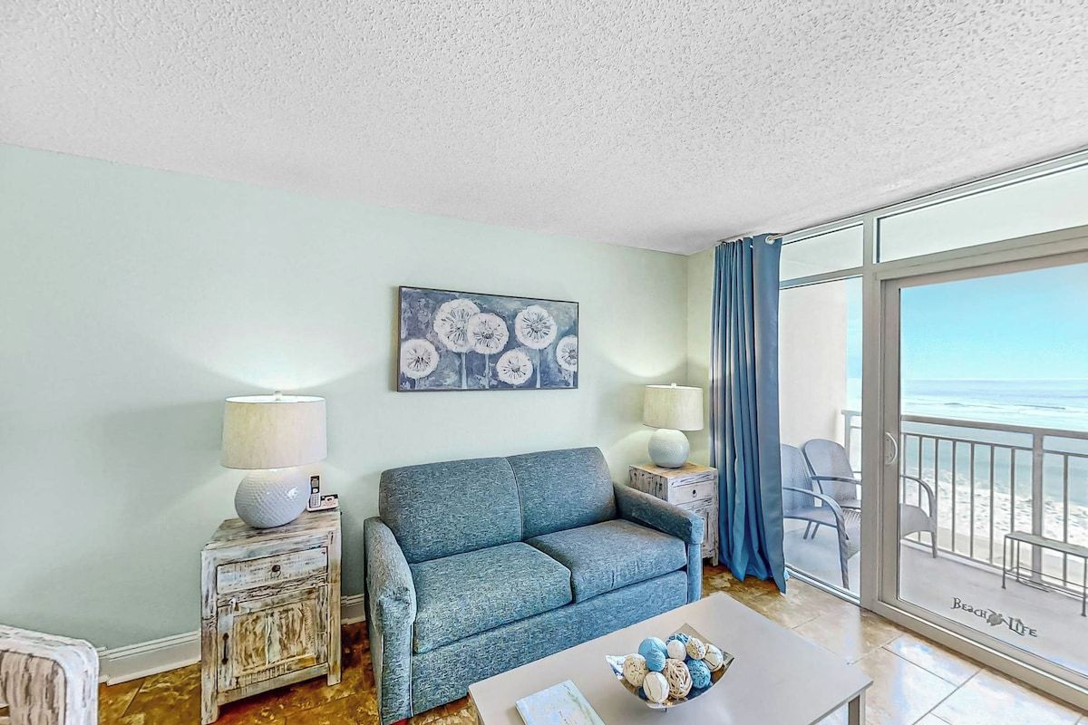 A living room with a sofa, a coffee table with decorative items, a painting on the wall, and a view of the beach through a large window.