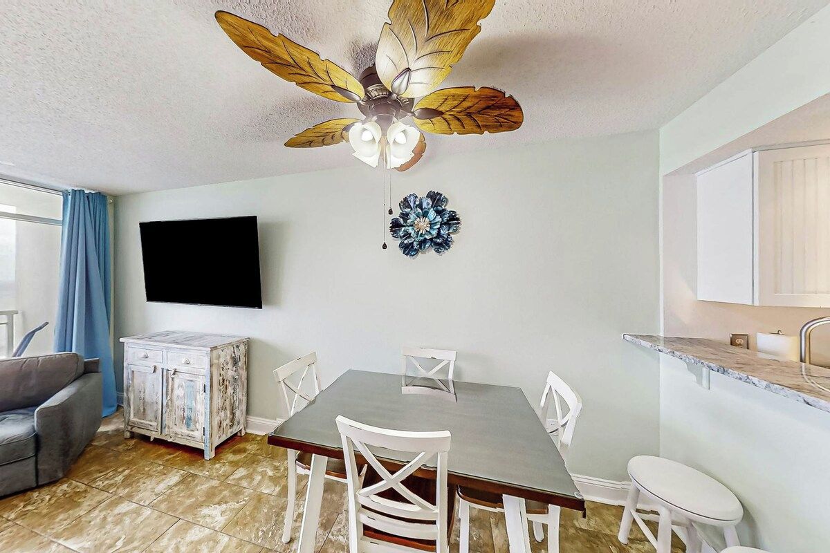 Un comedor con una mesa, sillas, ventilador de techo, televisor y un mueble de almacenamiento.