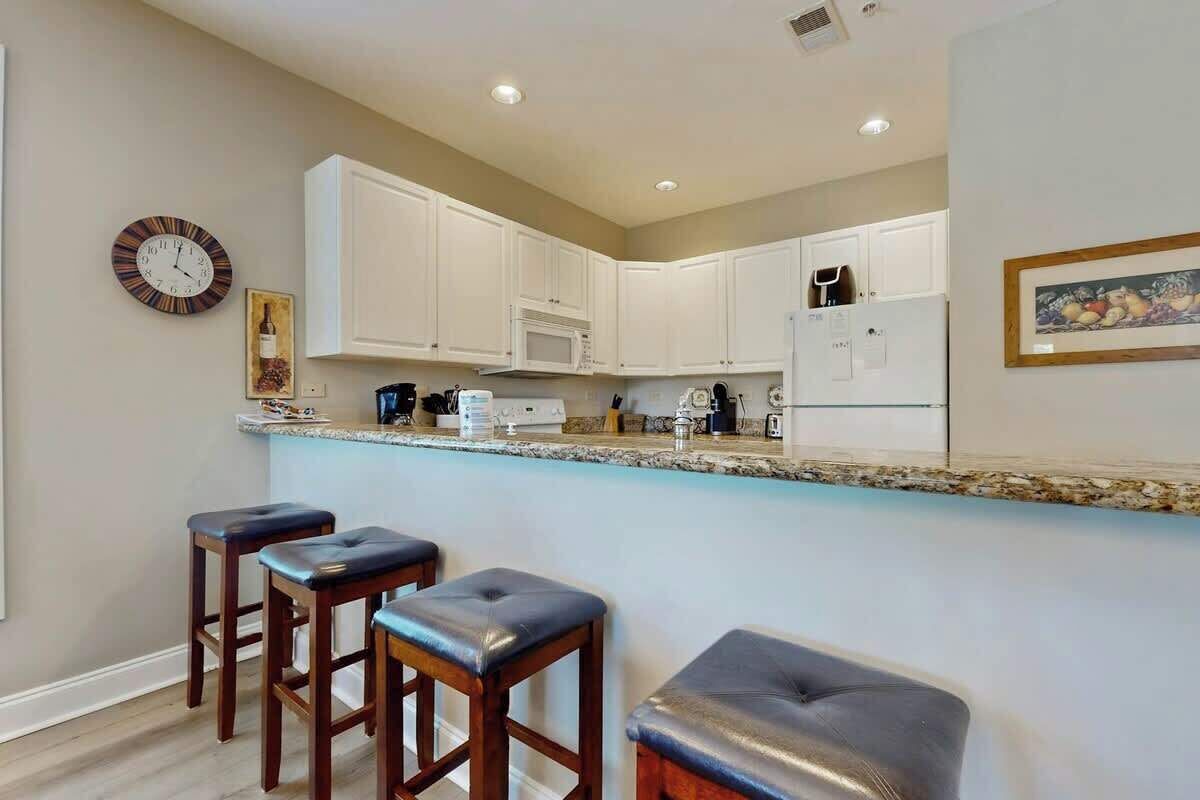 A kitchen with white cabinets, a microwave, and a refrigerator. There are three stools and a clock on the wall.