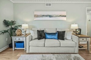 A living room with a sofa, two side tables, a lamp, a clock, a plant, and a painting on the wall. - Waipani Unit D-12 Condo (North Myrtle Beach)