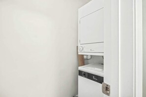 A compact, white laundry room with a built-in washer and dryer, a small shelf, and a door with a handle. - Waipani Unit D-12 Condo (North Myrtle Beach)