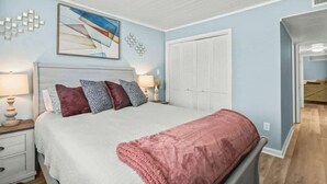 A bedroom with a bed, bedside tables, a nightstand, a window with blinds, and a door leading to another room. - Waipani Unit D-12 Condo (North Myrtle Beach)