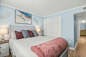 A bedroom with a bed, bedside tables, a nightstand, a window with blinds, and a door leading to another room. - Waipani Unit D-12 Condo (North Myrtle Beach)