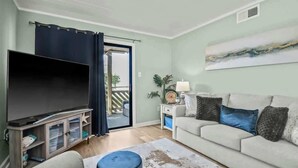 A living room with a flat-screen TV, a sofa, a coffee table, and a sliding glass door leading to an outdoor area. - Waipani Unit D-12 Condo (North Myrtle Beach)