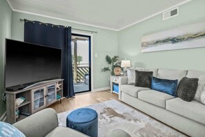 A living room with a flat-screen TV, a sofa, a coffee table, and a sliding glass door leading to an outdoor area. - Waipani Unit D-12 Condo (North Myrtle Beach)