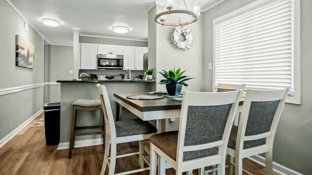 A modern kitchen with white cabinets, a central island, and a dining area with white chairs. - Waipani Unit D-12 Condo (North Myrtle Beach)