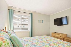 A bedroom with a bed, a television on the wall, a window with curtains, and a small piece of wall art. - Tropical Luxe | Waipani D-Ten (North Myrtle Beach)