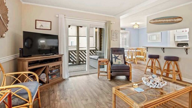 A living room with a television, wicker furniture, a glass coffee table, and a sliding glass door leading to a balcony. - Waipani Unit D-10 Condo (North Myrtle Beach)