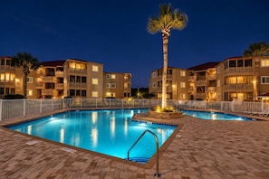 A swimming pool with a palm tree and a brick walkway, surrounded by multi-story buildings with balconies. - Tropical Luxe | Waipani D-Ten (North Myrtle Beach)