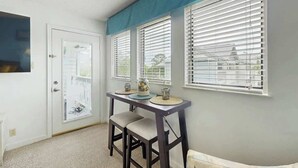 Um quarto com uma porta branca, uma pequena mesa com duas cadeiras e três janelas com persianas.