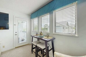 Ein Zimmer mit einer weißen Tür, einem kleinen Tisch mit zwei Stühlen und drei Fenstern mit Jalousien.