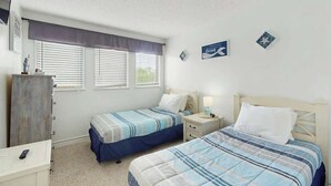 Quarto com duas camas, uma cômoda, um criado-mudo e uma janela com persianas.