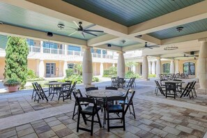 A covered outdoor patio area with multiple round tables and chairs, a large potted plant, and a view of a building with balconies in the background. - North Beach Villas 201 (North Myrtle Beach)