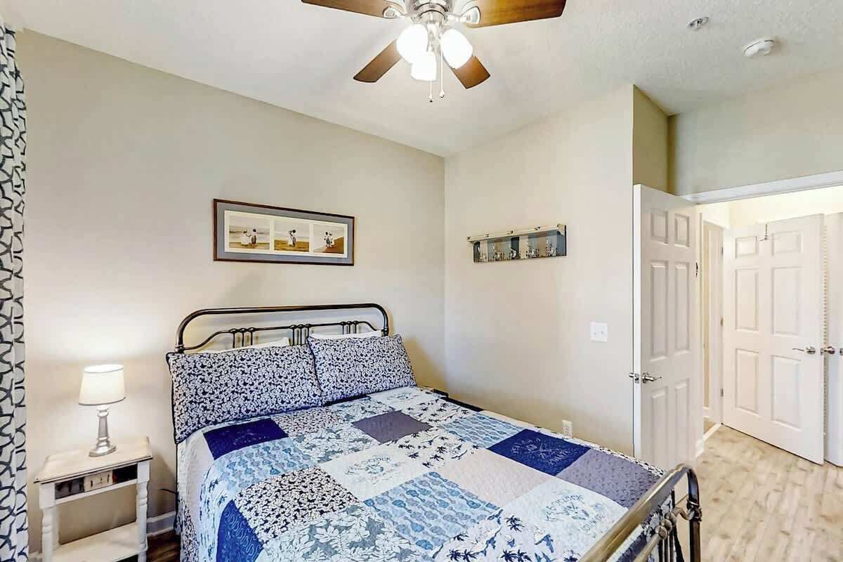 A bedroom with a bed, a ceiling fan, a bedside lamp, a framed picture on the wall, and a door.