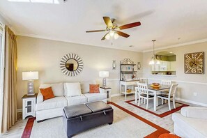 Ein Wohnzimmer mit Deckenventilator, ein Essbereich mit Tisch und Stühlen sowie eine Küche mit Schränken und einer Uhr.