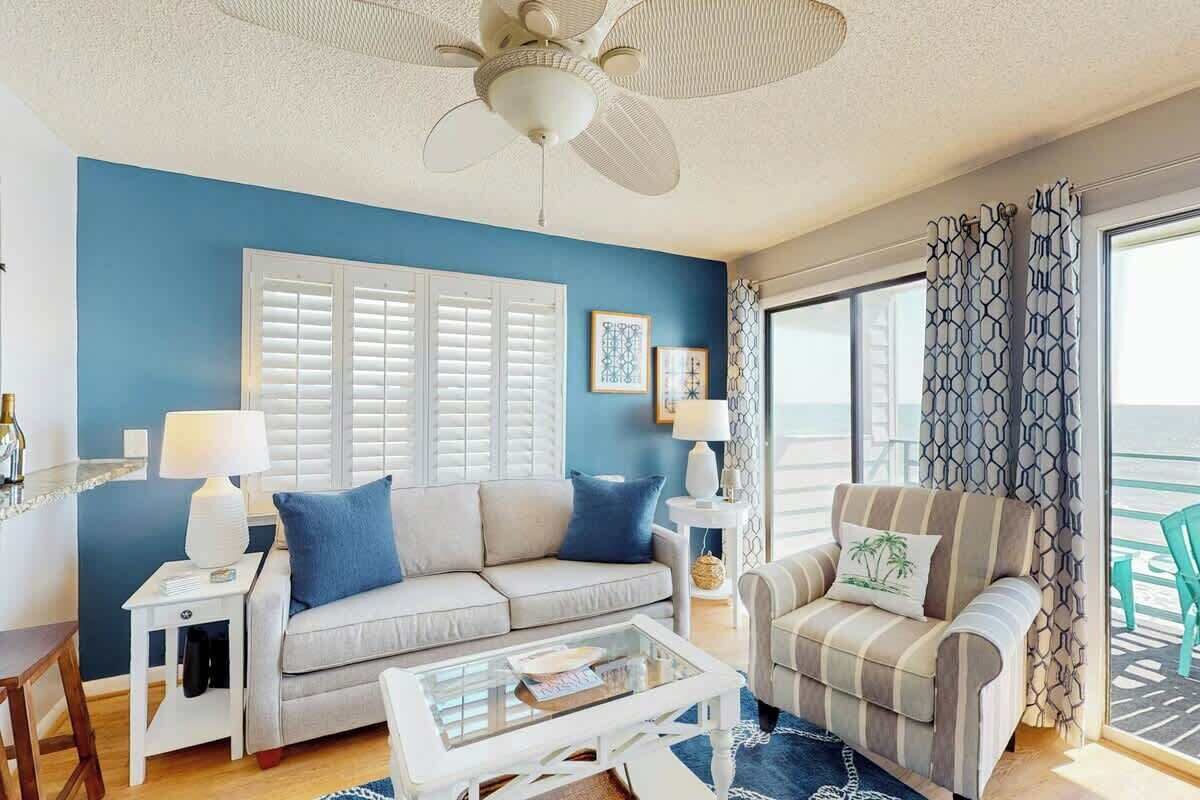 A living room with a blue wall, white shutters, a ceiling fan, and a view of the outdoors.