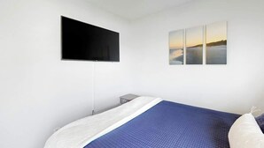A bedroom with a bed, a flat-screen TV mounted on the wall, and a set of three paintings depicting a beach scene. - Paradise Palms Retreat (Murrells Inlet)