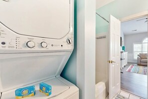 A laundry room with a white washing machine, two boxes of stain remover, and a door leading to a living area with a sofa and a window. - Gardens at Cypress Bay 10-302 (Little River)