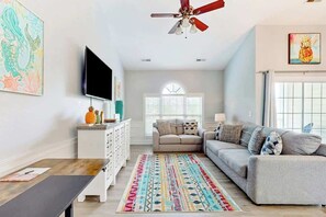 A living room with a sofa, TV, ceiling fan, and artwork. - Gardens at Cypress Bay 10-302 (Little River)