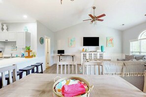A modern kitchen with white cabinetry, a dining area with a table and benches, a ceiling fan, and artwork on the walls. - Gardens at Cypress Bay 10-302 (Little River)