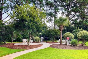 A landscaped outdoor area with a paved walkway, seating areas, and various trees. - Gardens at Cypress Bay 10-302 (Little River)
